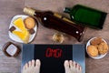 Female feet on digital scales with word omg on screen. Bottles and glasses of alcohol, plates with sweet food.