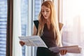 Female financial analyst holding papers studying documents standing against window with city view