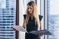 Female financial analyst holding papers studying documents standing against window with city view