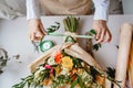 a female florist ties a green ribbon bow on a bouquet of flowers wrapped in craft paper on the desktop. Top view. Royalty Free Stock Photo