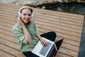 Female freelancer working on laptop, sitting on a pier by the backyard lake, headphones on, listening music. Concept of Royalty Free Stock Photo