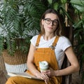 Female gardener in glasses resting, sitting on wooden chair in home greenhouse, hold collapsible silicone eco muge coffee/tea mug