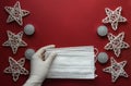 Female gloved hands holding medical masks with Christmas decorations on a red background. Copyspace. Flatlay