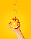 Female hand with bright manicure squeezes half of orange on yellow background. Pop art food photography. Drops of juice