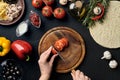 Female hand cut tomatoes on wooden board on kitchen table, around lie ingredients for pizza: vegetables, cheese and Royalty Free Stock Photo