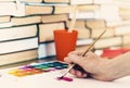 Female hand draws with a brush and watercolor paint on a sheet of paper on wooden table with stack of books background