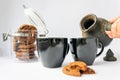 Female hand is going to pour aromatic coffee from dark clay cezve into black cups on a white background. Jar of chocolate chip