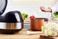 Female hand hold ladle over electric cooker in kitchen. Spoon with vegetables and modern multi cooker on wooden table. Royalty Free Stock Photo