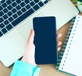 Female hand holding black mobile smart phone with blank screen on laptop computer and notepad background. Royalty Free Stock Photo