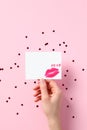 Female hand holding blank paper card mockup with lipstick kiss over pink background with confetti hearts. Happy Valentines Day or Royalty Free Stock Photo