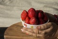 A female hand holding fresh organic red strawberry in a bowl on wooden table