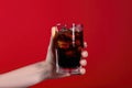 Female hand holding glass of cola with ice cubes on red background. Generative AI Royalty Free Stock Photo