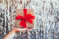 Female hand holding glittering golden gift box with velvet red ribbon with a bow on the sparkling gold tinsel background Royalty Free Stock Photo