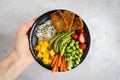 A female hand holding a homemade vegan poke bowl with tofu, mango, avocado on grey background