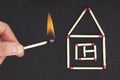 Female hand holding a lighted matchstick close to a flat house made of unused matches. Creative flat lay composition. Royalty Free Stock Photo