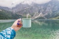 Female hand holding mint color enamel cup. Crystal clear lake and mountains. Tatra National park, Poland. Famous lake