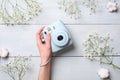 Female hand holding modern polaroid camera on a blue wooden background with flowers. Top view, tender minimal flat lay style compo