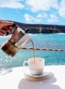 Female hand holding moka pot while pouring hot coffee into white cup on table against ocean and blue sky Royalty Free Stock Photo