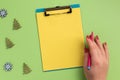 Female hand holding pen, clipboard with yellow sheet and christmas decor, on green background Royalty Free Stock Photo