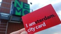 Female hand holding a red tourist guest card `I Amsterdam` on the background of signs of the Nemo Museum. Card for museums