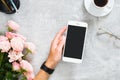 Female hand holding smartphone with blank screen mockup. Composition with roses flowers, coffee cup, stationery, glasses on Royalty Free Stock Photo