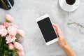 Female hand holding smartphone with blank screen mockup. Composition with roses flowers, coffee cup, stationery, glasses on Royalty Free Stock Photo