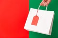 Female hand holding white blank paper shopping bag with price tag isolated on red and green background. Black friday Royalty Free Stock Photo