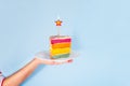 Female hand holding white plate with slice of Rainbow cake with birning candle in the shape of star isolated on blue background. Royalty Free Stock Photo