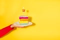 Female hand holding white plate with slice of Rainbow cake with birning candle in the shape of star isolated on yellow background Royalty Free Stock Photo