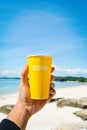 Female hand holding yellow cup with cool word sea and blue sky background