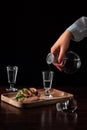 Female hand holds crystal decanter or carafe and pours on vodka in a glass. tasty appetizer bruschetta with bacon and pickles on