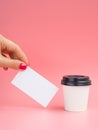 Female hand holds empty business card near a small paper coffee cup on a pink background. White espresso cup with blank Royalty Free Stock Photo