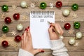 Female hand holds pen, writing Christmas shopping list, gift ideas on white notepad on wooden background Top view Flat lay Holiday Royalty Free Stock Photo