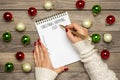 Female hand holds pen, writing Christmas shopping list, gift ideas on white notepad on wooden background Top view Flat lay Holiday Royalty Free Stock Photo