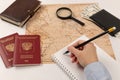 Female hand makes notes with a pencil in a notebook. on a white wooden table lies a map of the world, two passports, a magnifying