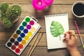 Female hand, palette with watercolors, brushes, cactus, cup of coffee, pencil, monstera leaf drawing on white paper on wooden tabl Royalty Free Stock Photo