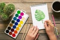 Female hand, palette with watercolors, brushes, cactus, cup of coffee, pencil, monstera leaf drawing on white paper on wooden tabl