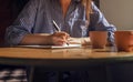 Female hand with pen taking notes, writing plans in diary on wood table with coffee cups in coffeehouse. Woman at wooden Royalty Free Stock Photo