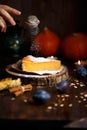 Female hand pouring a piece of pumpkin cheesecake with icing sugar, pumpkins, table lamp on a dark wooden background
