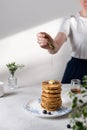Female hand pouring some honey or maple syrup on stack of pancakes with blueberries and piece of butter