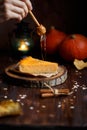 Female hand pours honey pumpkin cheesecake. Pumpkins, table lamp, foliage, vanilla on a wooden dark background. Autumn