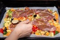 Female hand pours sauce for marinating pieces of pork on finely chopped and seasoned vegetables on a baking sheet for cooking in Royalty Free Stock Photo