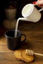 A female hand pours milk from a jug into a cup with rusks and honey beside it