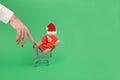 Female hand pull a grocery trolley shopping cart with gift with a red ribbon wearing Santa hat in on a green background Royalty Free Stock Photo