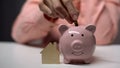 Female hand putting coin in piggybank, wooden toy home standing near, mortgage Royalty Free Stock Photo