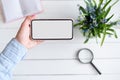 Female hand with a smartphone. White blank screen. Table with notebook and flowers on background Royalty Free Stock Photo