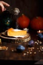 Female hand sprinkles powdered sugar on pumpkin cheesecake. Pumpkins, table lamp, foliage, vanilla on a wooden dark background.
