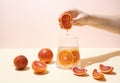 Female hand squeezing half of juicy blood orange into a glass of functional water with citrus slices on a yellow-pink background