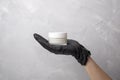 Female hands in black medical glove holds jar with cream. Women`s hands with white cosmetic jar on the grey background