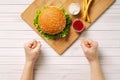 Female hands clenched into fists, on a wooden board lies a hamburger and french fries. Manifestation of willpower. Light wooden